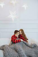 jovem lindo casado casal dentro Coincidindo vermelho pijamas sentado em uma cinzento sofá, embrulhado dentro uma cobertor, abraçando e se beijando. Novo anos feriados e presentes debaixo a Natal árvore foto