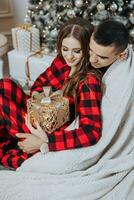 jovem lindo família casal dentro idêntico vermelho pijamas perto a Natal árvore, embrulhado dentro uma cobertor, alegrar e troca Novo anos presentes. Novo anos feriados e presentes debaixo a Natal árvore foto