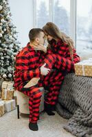 uma jovem lindo casado casal dentro a mesmo vermelho pijamas, perto a Natal árvore, estão sorridente, abraçando e se beijando. Novo ano feriados e presentes debaixo a Natal árvore foto