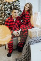 uma jovem lindo família casal dentro a mesmo vermelho pijamas, perto a Natal árvore, alegrar e troca Novo anos presentes. Novo ano feriados e presentes debaixo a Natal árvore foto