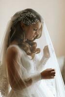 retrato do uma Loiras noiva dentro uma Casamento vestir com mangas debaixo uma véu, em pé dentro perfil dentro dela quarto foto