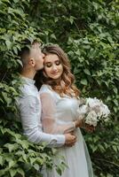 noiva e noivo ficar de pé em uma fundo do verde folhas. a noivo Beijos a noiva em a têmpora foto