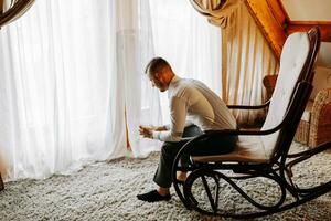 uma jovem homem de negocios dentro uma hotel quarto senta em uma cadeira e bebidas dele uísque a partir de uma vidro. descansar dentro Maravilhoso apartamentos. do noivo manhã às a hotel foto
