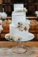 uma branco de dois níveis Casamento bolo, decorado com branco flores e cinza por aí a arestas, carrinhos em uma ficar de pé perto uma espelho. Casamento sobremesas. foto