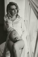 estúdio retrato, fechar-se, do uma lindo grávida jovem mulher em uma branco cama, segurando dela grávida barriga com dela mãos foto