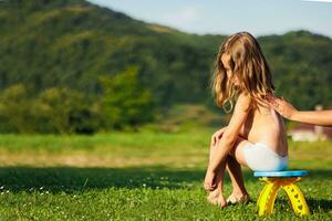 uma mãe aplica-se protetor solar para dela pequeno da filha costas antes a criança vai para uma andar. a criança é sentado em uma cadeira e verde grama. livre espaço foto