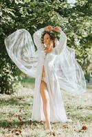 retrato do uma sonhadores noiva em pé dentro uma jardim. a lindo noiva é vestido dentro uma branco roupão e uma tiara do fresco flores em dela cabeça. foto