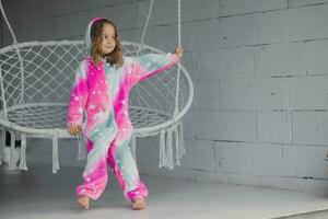 retrato do feliz pequeno menina em varanda sentado em vime balanço e jogando dentro Rosa pijamas foto