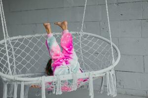 feliz pequeno menina em a varanda sentado em uma vime balanço e jogando dentro Rosa pijamas, levanta dela pernas para a topo foto