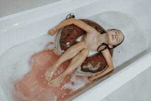pequeno, sorridente, lindo cabelos escuros menina com grandes cabelo, criança banha, lavagens dentro uma branco banho com espuma. Diversão foto. foto