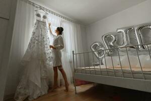 uma lindo jovem noiva dentro uma branco anágua é em pé de a janela, tocante dela Casamento vestir. final preparativos para a casamento. a noiva é esperando para a noivo. manhã, noiva. foto