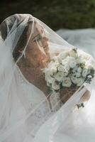 uma noiva dentro uma Casamento vestir com uma grandes trem e uma véu detém uma Casamento ramalhete do rosas enquanto sentado debaixo uma véu em verde grama. foto a partir de acima