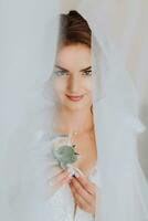 a noiva é vestido dentro a elegante vestir, coberto com uma véu, posando e segurando uma flor na lapela. Casamento foto, manhã do a noiva foto