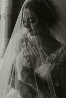 a noiva é vestido dentro a elegante vestir, coberto com uma véu, posando e segurando uma flor na lapela. Casamento Preto e branco foto, manhã do a noiva foto