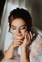 a manhã do a noiva dentro uma branco roupão e a elegante Penteado. a adulto menina senta em uma cadeira e aperta a brinco, olhando para dentro a Câmera lente e sorridente foto