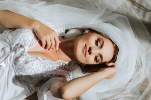 uma lindo jovem noiva dentro uma branco roupão poses para uma fotógrafo, deitado em a chão, em uma véu. Casamento fotografia, fechar-se retrato, chique Penteado. foto