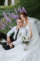 retrato do a noiva e noivo dentro a parque. a noiva é segurando uma ramalhete e abraçando dela noivo, elas estão sentado, posando perto lavanda flores Casamento andar dentro a parque. grandes trem do a vestir foto