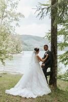 uma Casamento casal é desfrutando a melhor dia do seus vidas contra a pano de fundo do uma lago e alta árvores a noivo abraços a noiva. foto