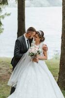 uma Casamento casal é desfrutando a melhor dia do seus vidas contra a pano de fundo do uma lago e alta árvores a noivo abraços a noiva. foto