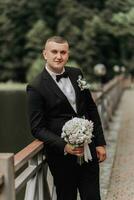 Casamento retrato. uma homem dentro uma Preto terno carrinhos em uma ponte com uma ramalhete e parece ausente. noiva. masculino, o negócio estilo foto