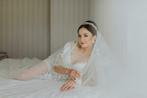 uma lindo menina com uma Casamento Penteado e uma tiara em dela cabeça dentro uma transparente manto, deitado em uma cama dentro uma hotel com uma real interior. grandes véu, natural luz, retrato foto