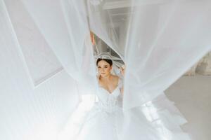 preparação para a casamento. lindo jovem noiva dentro branco luxuoso Casamento vestir, tiara em cabeça, grandes véu dentro real hotel sala. luxo sorridente modelo foto