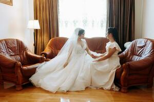Casamento tema. a noiva dentro uma branco vestir é sentado com dela amigo em a sofá. a último minutos do preparativos para a casamento. foto