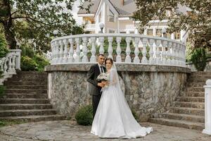 retrato do uma jovem noiva dentro uma branco vestir e uma morena noivo dentro uma terno em a escadas abraçando perto a velho cidade parque. uma lindo e romântico casamento, uma feliz casal foto