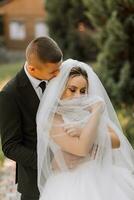 uma à moda noivo dentro uma Preto terno e uma fofa noiva dentro uma branco vestir com uma grandes véu estão abraçando dentro uma parque. Casamento retrato do sorridente e feliz recém-casados. foto