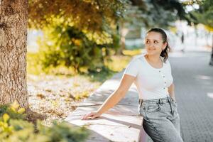 uma lindo menina dentro uma branco suéter e jeans é em repouso dentro a cidade. conceito do trabalhos e descansar. bastante a ternura do a modelo, olhando para a lado. foto