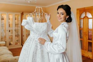 feliz morena noiva dentro branco cetim vestir com grandes véu, profissional Maquiagem e Penteado. a noiva é em pé dentro a quarto Próximo para dela Casamento vestir, que é em uma manequim. foto