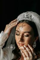 lindo noiva com grandes Sombrio encaracolado cabelo dentro uma grandes branco roupão dentro a manhã. a noiva é preparando para a casamento. profissional Maquiagem e cabelo. uma coroa em a cabeça e uma grandes véu do a noiva foto