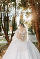 lindo noiva debaixo uma véu dentro uma elegante Casamento vestir em uma natural fundo dentro a parque às pôr do sol. uma deslumbrante jovem noiva é incrivelmente feliz. feliz menina em dela Casamento dia foto