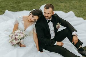 elegante noivo e fofa morena noiva dentro branco vestir, inclinado em do noivo ombro, rindo dentro parque sentado em grama, jardim, ao ar livre. Casamento fotografia, retrato do sorridente recém-casados. foto