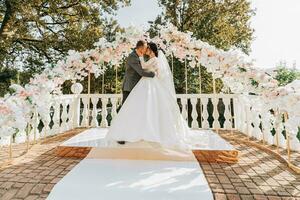 feliz noiva e noivo se beijando às a Casamento cerimônia. a Casamento arco é decorado com rosa flores foto