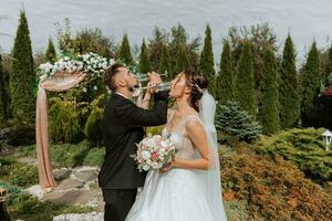 feliz noiva e noivo às a Casamento cerimônia bebendo champanhe. a Casamento arco dentro a jardim é decorado com rosa flores foto