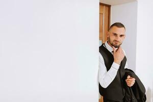 retrato do uma homem dentro uma branco camisa, colete e Preto gravata dentro uma quarto com natural claro. a noivo é preparando para a casamento. a homem é vestindo uma branco camisa. à moda noivo foto