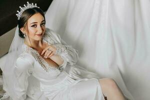 a incrivelmente lindo noiva dentro a manhã dentro uma cetim roupão com uma tiara em dela cabeça poses Próximo para dela Casamento vestir foto