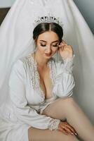 a incrivelmente lindo noiva dentro a manhã dentro uma cetim roupão com uma tiara em dela cabeça poses Próximo para dela Casamento vestir foto