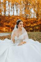 uma lindo noiva é sentado em a Relva perto a outono folhas do a árvores Casamento retrato do uma jovem noiva dentro a outono tema. Casamento fotografia. foto