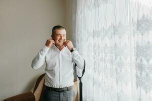 retrato do uma jovem sorridente homem dentro uma camisa dentro dele sala. a noivo pega vestido e prepara para a Casamento cerimônia foto