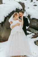 Casamento dentro inverno. a noiva e noivo estão em pé contra a fundo do coberto de neve pedras. a noiva dentro uma branco vestir e branco poncho. noivo dentro uma Preto casaco foto