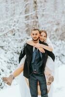 lindo Casamento casal caminhando dentro inverno Nevado floresta, mulher dentro branco vestir e vison pele casaco, barbudo homem dentro Preto casaco. a noivo carrega a noiva dentro dele braços, uma engraçado foto