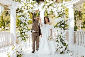 sensível cerimônia do a noiva e noivo. uma feliz recém-casado casal carrinhos contra a fundo do uma Casamento arco decorado com fresco flores a noiva e noivo mostrar fora seus Casamento argolas foto