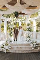 sensível cerimônia do a noiva e noivo. uma feliz recém-casado casal carrinhos contra a fundo do uma Casamento arco decorado com fresco flores foto