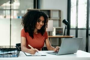 jovem mulher digitando em tábua e computador portátil enquanto sentado às a trabalhando dentro escritório foto