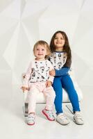 dois à moda pequeno meninas sentar em 1 volta cadeira em branco parede fundo foto