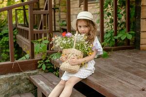 fofa pequeno menina sentado em porta do país casa com cesta do flores silvestres foto