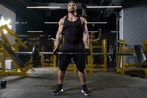 barbudo atleta dentro Preto esporte luvas, shorts, colete, boné e tênis. ele elevação uma barra, posando dentro Sombrio Academia com amarelo equipamento. cheio comprimento foto