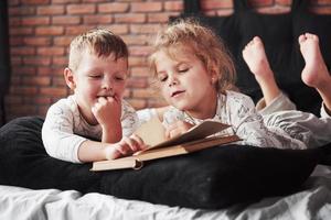 duas crianças deitam-se em uma cama grande e lêem um livro interessante. eles estão vestidos com o mesmo pijama foto